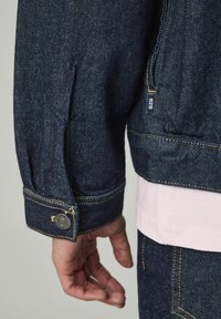 Veste en denim foncé avec coutures jaunes, poignets boutonnés, et une petite étiquette de marque près de l'ourlet. Portée sur une chemise rose clair.