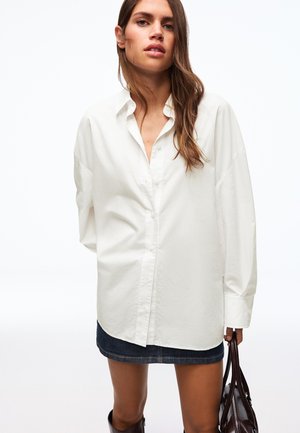 Jonge vrouw met lang bruin haar die een oversized wit overhemd draagt, een donker denim rok en een donkerbruine leren tas vasthoudt.