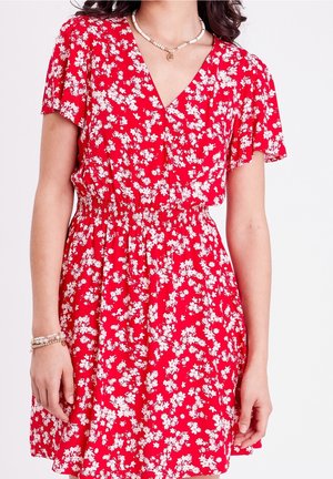 Femme portant une robe rouge avec des motifs floraux blancs, manches courtes, encolure en V et taille élastique, accessoirisée avec des bracelets et un collier en coquillage.