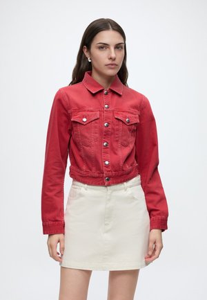 Femme portant une veste en jean rouge boutonnée et une jupe blanche, debout sur un fond blanc uni.