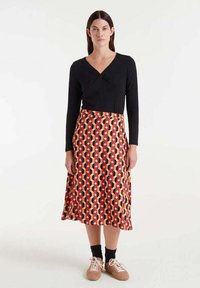 Top negro de manga larga con un diseño cruzado combinado con una falda de patrón colorido que presenta formas rojas, naranjas y negras. Se llevan zapatos marrones.