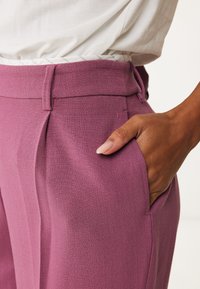 Pruimenkleurige op maat gemaakte broek met een gladde textuur, voorplooien en zijzakken. Een hand rust casual in de zak.