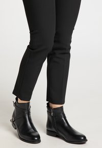 Botine negre până la gleznă, cu fermoare laterale și curele decorative, purtate cu pantaloni negri slim-fit pe un fundal simplu.