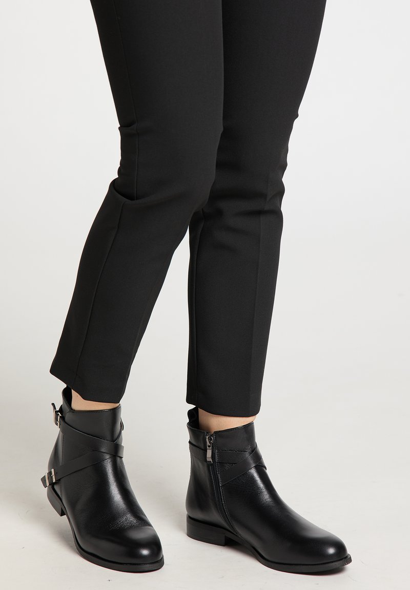 Botine negre până la gleznă, cu fermoare laterale și curele decorative, purtate cu pantaloni negri slim-fit pe un fundal simplu.