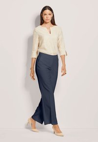 Vrouw in een crèmekleurige, gestructureerde blouse met driekwartmouwen, marineblauwe, wijde broek, beige hakken en een gouden armband die loopt tegen een lichte achtergrond.