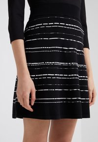 Schwarzes Kleid aus strukturiertem Stoff, mit horizontalen weißen Streifen unterschiedlicher Längen. Der Rock hat eine ausgestellte Form und einen taillierten Bund.