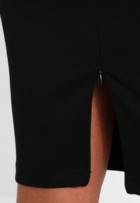 Zwarte aansluitende rok gemaakt van een rekbare stof, voorzien van een zijrits en een split voor meer bewegingsvrijheid. Zachte textuur, gladde naden.