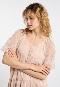 Světle růžová blůza s krátkými rukávy, složitým paisley vzorem, nařaseným výstřihem a volným střihem, která má jemnou, splývavou texturu látky.