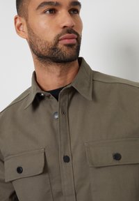 Olijfgroene shirtjas met een puntige kraag, twee borstzakken, zwarte drukknopen en een minimalistisch ontwerp, gemaakt van duurzaam materiaal.