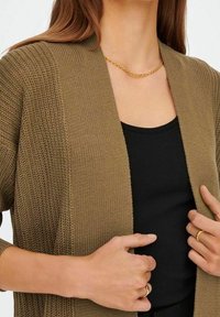 Cardigan en tricot vert olive avec un design à ouverture devant. Débardeur noir en dessous, colliers en chaîne dorée et bagues aux doigts.