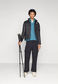 Crna prozračna jakna preko plavog flisa sa patentnim zatvaračem, u kombinaciji s crnim cargo hlačama. Drži treking štapove, nosi cipelice boje kreme.