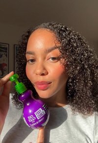 Bouteille de produit pour cheveux violette avec un dessus de pompe vert, étiquetée "Bed Head Foxy Curls." Le sujet présente des cheveux bouclés et porte des vêtements gris clair.