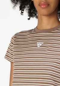 Kahvipunainen raidallinen t-paita lyhyillä hihoilla, ribatulla kauluksella ja pienellä kolmiomaisella "GUESS"-logopatchilla rinnassa. Kangas näyttää pehmeältä.