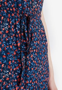 Gros plan d'une robe bleu marine avec un motif floral rouge et bleu et une ceinture en tissu nouée à la taille, portée par une personne avec un bras nu visible.