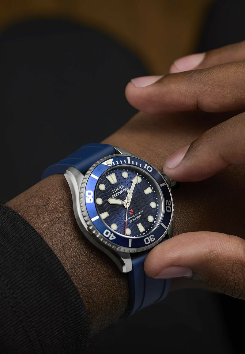 Zegarek Timex Deepwater z niebieskim kauczukowym paskiem i stalową obudową. Tarcza wyróżnia się wzorem fal, luminescencyjnymi znacznikami oraz jednostronnie obracanym pierścieniem.