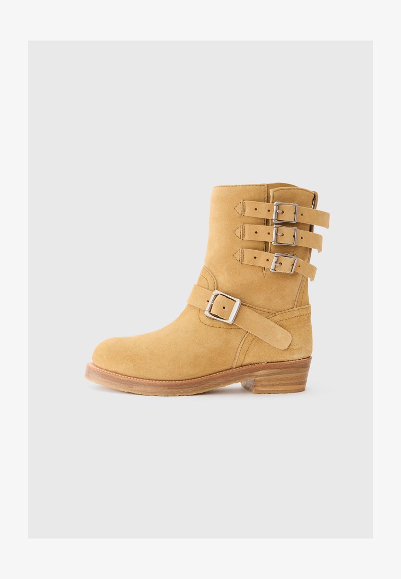 Keltainen mokkanahkainen nilkkabootsi, jossa on pyöreä kärki, kolme säädettävää hopeista solkivöitä ja matala pinottu puinen korko. Silkkinen pinta.