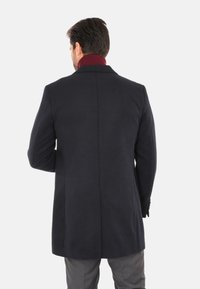 Cappotto in lana di un blu scuro, con taglio dritto, caratterizzato da un alto colletto bordeaux e dettagli con bottoni sui polsini. Texture liscia e design minimalista.