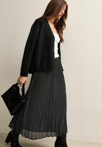 Black cropped cardigan over a white tank, paired with a long black polka dot pleated skirt and black knee-high boots, carrying a black bag.