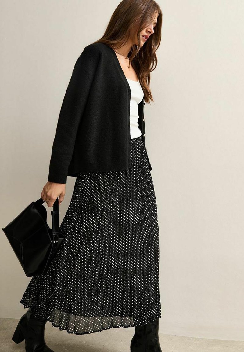 Black cropped cardigan over a white tank, paired with a long black polka dot pleated skirt and black knee-high boots, carrying a black bag.