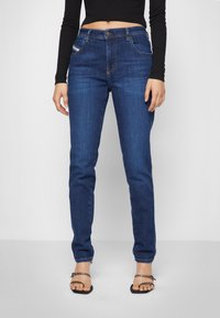 Jeans de denim azul escuro com um corte slim, apresentando cinco bolsos e costura laranja. A cintura possui um botão de metal e um fecho de zip.