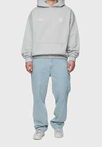 Sudadera gris con bolsillo frontal y logo en el pecho, combinada con jeans de corte relajado en azul claro con bordado rosa, y zapatillas grises.