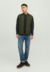 Grüne gepolsterte Weste mit vertikalen Nähten über einem navyblauen strukturierten Pullover, kombiniert mit hellblauen Jeans und schwarzen Schuhen mit einer sandfarbenen Sohle.