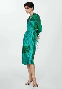 Rochie verde, cu mâneci lungi, cu modele geometrice și un detaliu cu funda în talie. Material neted, ușor, cu o finisare lucioasă.