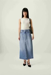 Woman wearing sleeveless white top, long faded blue denim skirt, black heels, and carrying black shoulder bag, standing on plain background.