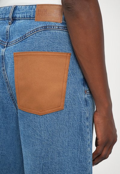 Blå denimjeans har en stor, beige läderlappficka på baksidan. Ytan är slät, med synliga sömdetaljer.
