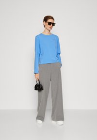 Maglia azzurra chiara a maniche lunghe con scollatura rotonda, abbinata a pantaloni grigi a gamba larga. Gli accessori includono occhiali da sole neri oversize e una mini borsa.