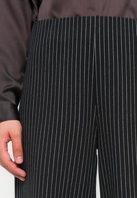 Crne hlače s prugicama u bijeloj boji, s vertikalnim linijama, koje nude prilagođeni kroj i glatku tkaninu. Ruka počiva na nogavici hlača.