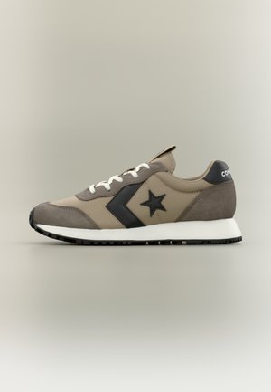 Bež in sivo supergo Converse z črnim logotipom zvezde, belimi vezalkami in črnim podplatom na nevtralno sivi podlagi.