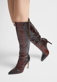 Botas de cuero marrón hasta la rodilla con textura de cocodrilo, punta afilada, tacón de aguja y detalle de cremallera lateral. Acabado suave con brillo lustroso.