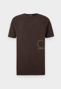 T-shirt de manga curta castanho escuro com um pequeno rosto contornado a ouro, usando óculos de sol e um rabo de cavalo, rotulado "KARL LAGERFELD" na parte da frente.