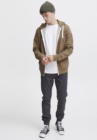 Bruine hoodie met rits over een wit T-shirt, gecombineerd met donkere broeken en zwarte sneakers. Het model draagt een gebreide beanie. Eenvoudige, casual look.