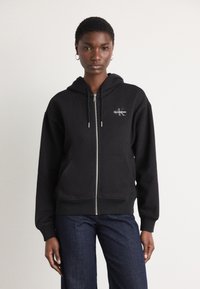 Black zip-up hoodie made of soft fabric with a front pocket, featuring a small white logo on the left chest and drawstring hood.