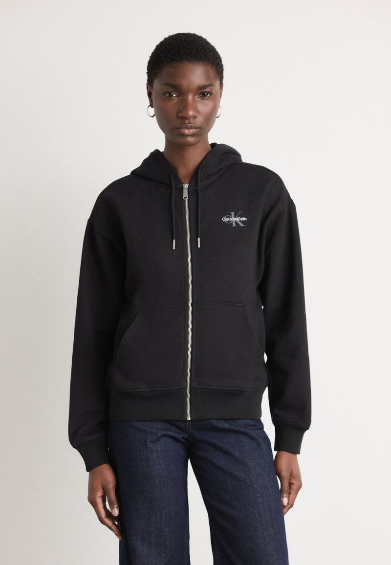 Schwarze Zip-Hoodie aus weichem Stoff mit einer vorderen Tasche, versehen mit einem kleinen weißen Logo auf der linken Brust und einer Kapuze mit Kordelzug.