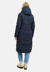 Navy puffertjacka med huva, med en lång design och chevron-sömmar. Kombinerad med ljusblå jeans och beiga ankelskor.