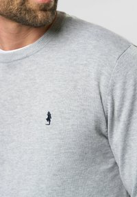 Grijze gebreide trui met een klassieke ronde hals. Opmerkelijke detail: klein geborduurd logo van een wandelende figuur op de linkerborst.