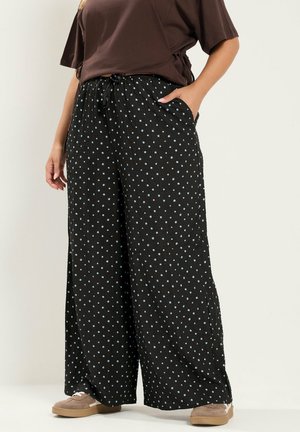 Persoon die zwarte broek met wijde pijpen met kleine blauwe stippen draagt, een bruin kortemouwtopje en beige casual schoenen, staat met één hand in de zak.