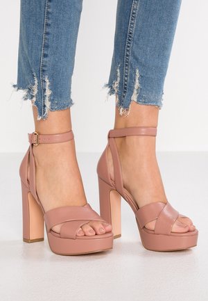 Pieds portant des sandales à plateforme à talons hauts rose poudré avec des brides autour de la cheville, associées à un jean skinny bleu à ourlet effiloché.