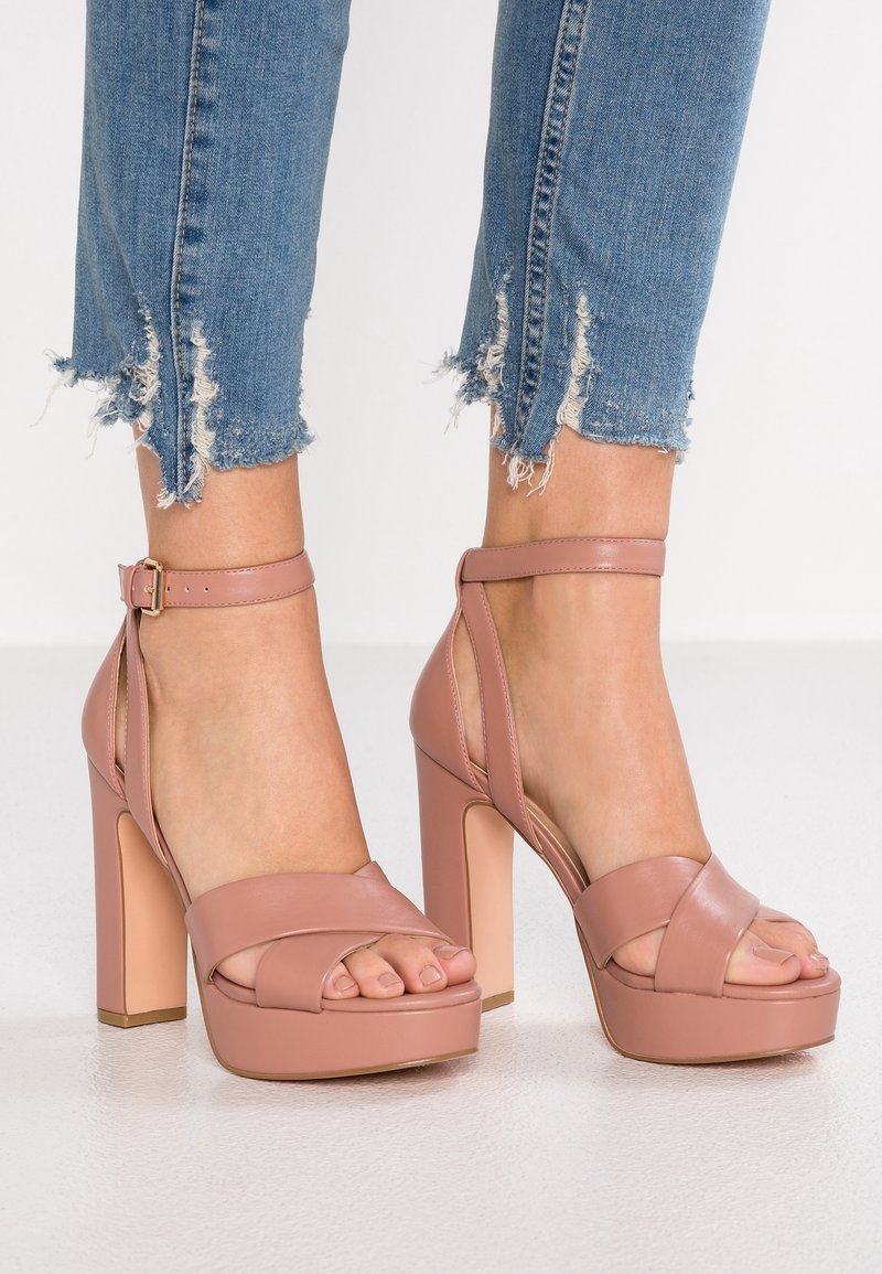 Pieds portant des sandales à plateforme à talons hauts rose poudré avec des brides autour de la cheville, associées à un jean skinny bleu à ourlet effiloché.