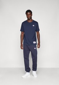 Camiseta de algodón azul marino con logo en el pecho, combinada con pantalones deportivos azul marino con rayas laterales y acentos de logo. Zapatillas blancas.