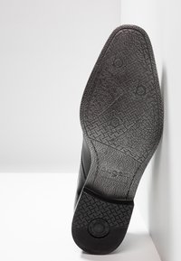 Semelle de chaussure en caoutchouc noir avec surface texturée, présentant une forme profilée et un logo de marque en relief. Grip à motif pour une meilleure traction.