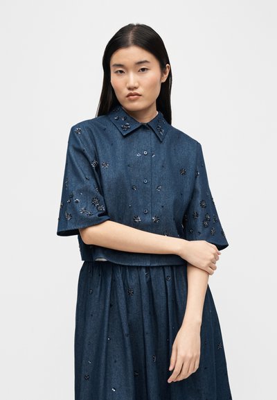 Jeune femme aux longs cheveux noirs portant une chemise boutonnée à manches courtes bleu foncé et une jupe assortie décorée de petites appliques florales noires.