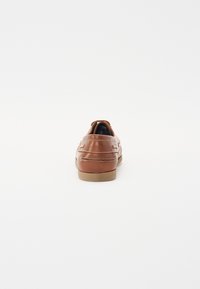 Zapato de cuero marrón con una textura suave, forma redondeada, detalle de costura blanca y una suela de goma en color marrón claro. Vista trasera.