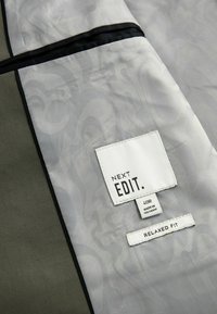 Grijze jas interieur met een subtiel patroon, zwarte afwerking en een label dat het merk, de maat en pasvormdetails aangeeft. Stof lijkt glad en duurzaam.