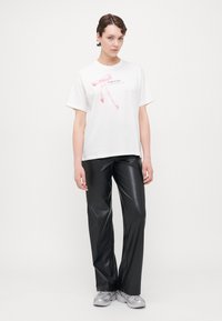 Vit bomulls T-shirt med rosa bandgrafik, parrade med svarta konstläderbyxor. Modellen bär gråa träningsskor.