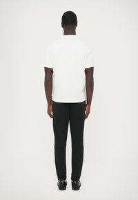 Balts kokvilnas t-krekls ar īsām piedurknēm, redzams no aizmugures, kombinācijā ar melnām sporta biksēm ar šauru piegriezumu un ribota auduma manžetēm.