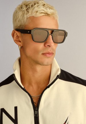 Plavokosi muškarac s kratkom kosom u velikim crnim sunčanim naočalama, malom zlatnom obručastom naušnicom, višeslojnim lancima i bijelom jaknom na patent s crnim detaljima.